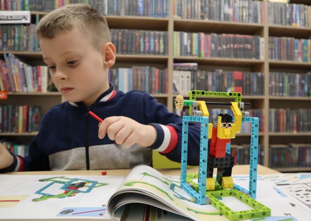 Na zdjęciu znajduje się chłopiec z klockiem w ręce przeglądający instrukcje do zbudowania stojącej po prawej stronie konstrukcji z LEGO przedstawiającą ludzika na drążku. W tle znajdują się półki z książkami.