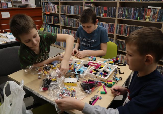 Na zdjęciu znajduje się trzech chłopców przy stoliku, na którym leżą klocki LEGO, z których chłopcy budują samochód. Jeden chłopiec wyciąga rękę aby sięgnąć klocek. W tle znajdują się półki z książkami.