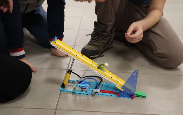 Na zdjęciu znajduje się skocznia narciarska z LEGO po której jedzie ludzik. W tle dookoła znajdują się nogi chłopców.