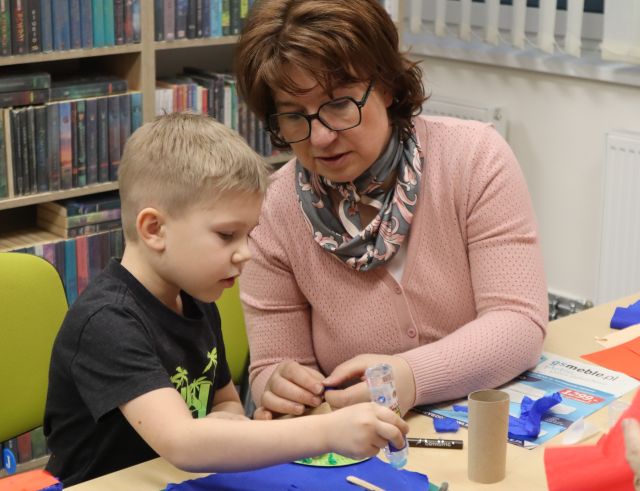 Na zdjęciu znajduje się kobieta, która pomaga chłopcu obok siebie, przed nimi leżą kolorowe kartki oraz rolka po papierze, a w tle znajdują się półki z książkami.