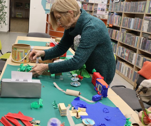 Na zdjęciu znajduje się kobieta nachylająca się nad znajdującą się na stole makietą, do której dokleja zielony element. Makieta przedstawia jezioro, bibliotekę, fontannę oraz drzewa. W tle na stoliku leży okrągły pojemnik, zielona kartka oraz kolorowe kartki. W tle znajdują się półki z książkami.