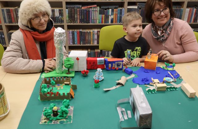 Na zdjęciu znajdują się dwie kobiety oraz jeden chłopiec między nimi, a przed nimi na stole znajduje się makieta miasta z biblioteką, drzewami, jeziorem i wieżą ciśnień. W tle znajdują się półki z książkami.