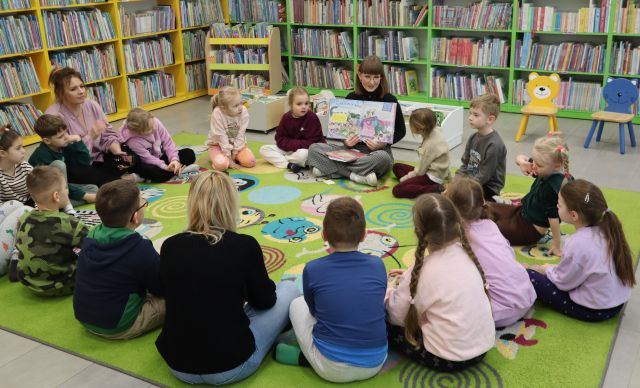 Na zdjęciu znajdują się przedszkolaki wraz z dwoma Paniami na dywanie w okręgu między nimi siedzi kobieta i pokazuje im ilustracje w książce. W tle znajdują się półki z książkami.