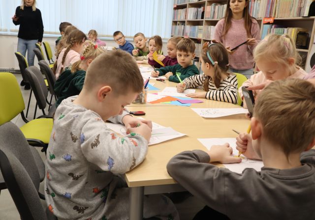 Na zdjęciu znajdują się przedszkolaki przy złączonych stołach i kolorują maski przed sobą, na stole znajdują się również kolorowe kartki , a za przedszkolakami znajdują się dwie kobiety. W tle znajdują się okna oraz półki z książkami po prawej stronie.