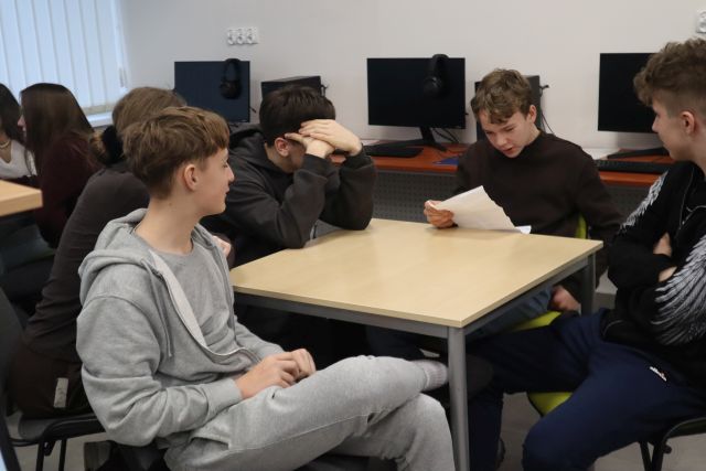 Na zdjęciu znajduje się grupka młodzieży przy stole, jeden chłopak trzyma w ręku kartkę, z której czyta napisaną przez grupę historię, w tle znajdują się komputery.