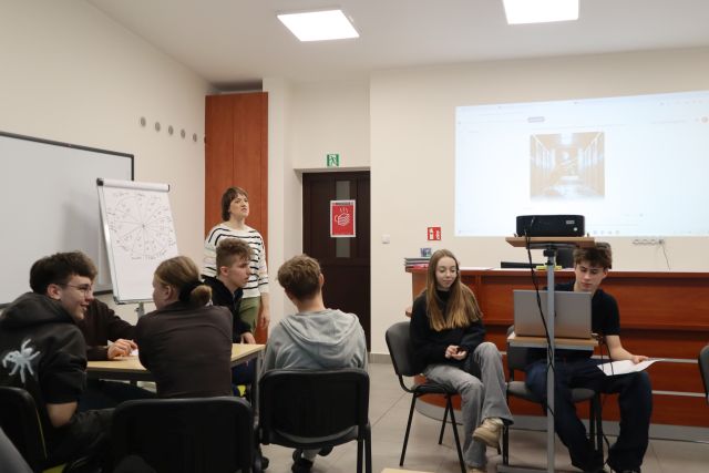Na zdjęciu po lewej znajduje się młodzież przy stolikach, za nimi stoi kobieta, a obok niej tablica z kartkami. Po prawej stronie znajduje się dwójka osób przy laptopie i za pomocą rzutnika wyświetlają tworzone przez AI obrazy pasujące do ich opowieści.