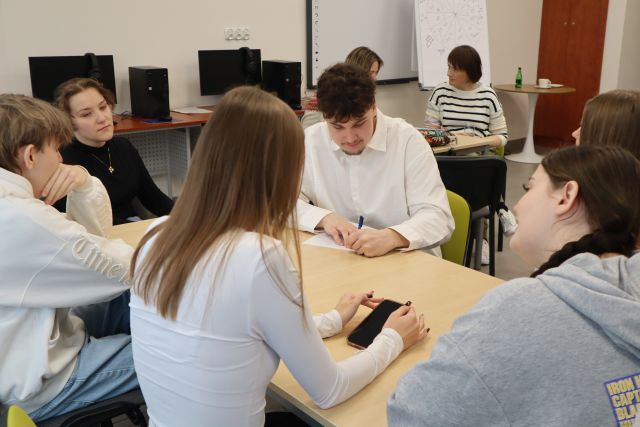 Na zdjęciu znajduje się szóstka młodzieży siedząca przy złączonych stołach , jedna dziewczyna ma przed sobą telefon, a chłopak na przeciwko niej pisze na kartce swoją część historii. W tle przy osobnym stoliku siedzą dwie kobiety. Warsztaty odbywają się w sali komputerowej.