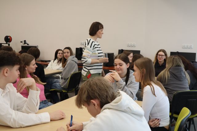 Na zdjęciu znajduje się młodzież przy stolikach, a między stolikami na środku stoi kobieta. Warsztaty odbywają się w sali komputerowej.