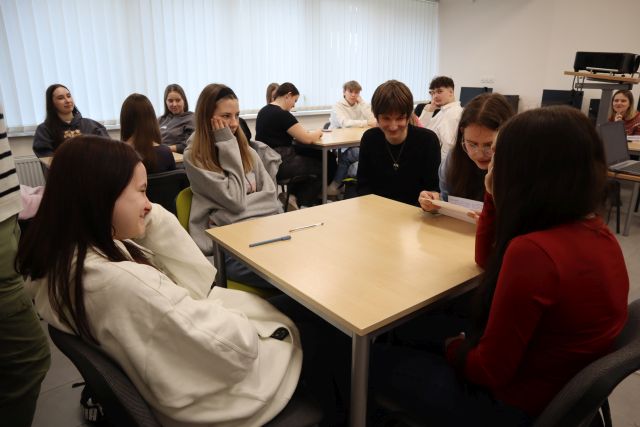 Na zdjęciu znajduje się młodzież przy stolikach, w centrum znajduje się stolik z piątką młodzieży, jedna z nich czyta historię zapisaną na kartce. W tle znajdują się okna.