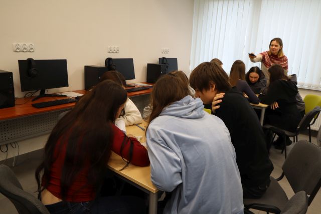 Na zdjęciu znajduje się młodzież przy stolikach, która pisze swoją historię, nad stolikiem z tyłu stoi kobieta i robi zdjęcie telefonem. Warsztaty odbywają się w sali komputerowej.