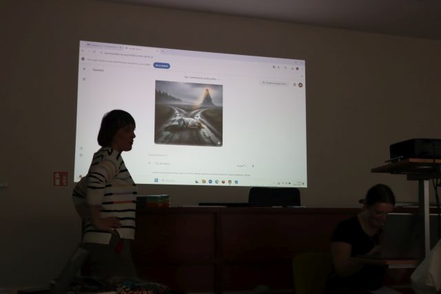 Na zdjęciu znajduje się obraz wyświetlany przez rzutnik na ścianie przedstawiający górę, na której znajduje się prezent, a przed górą leży załamany mężczyzna. Przed laptopem, który znajduje się pod rzutnikiem siedzi dziewczyna, a po lewej stronie stoi kobieta bokiem i ma ręce oparte na biodrach.