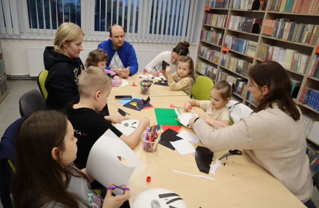Na zdjęciu przy złączonych stołach siedzi piątka dzieci oraz czworo rodziców, którzy wspólnie z dziećmi tworzą ze znajdujących się na stoliku kartek zebrę na papierowym talerzyku. W tle znajdują się okna, a po prawej stronie półki z książkami.