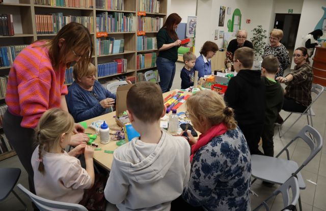 Na zdjęciu znajdują się dzieci oraz dorośli, którzy wspólnie tworzą makietę miasta przy złączonych stołach. W tle znajdują się półki z książkami.