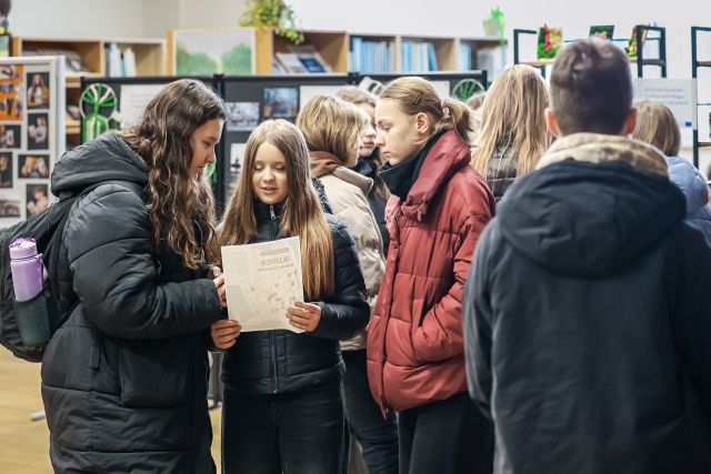 Na zdjęciu znajduje się grupka młodzieży, jedna dziewczyna trzyma przed sobą kartkę. W tle znajdują się półki z książkami.