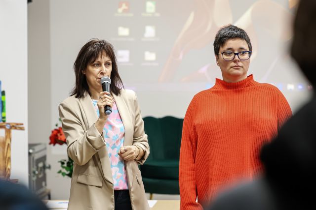 Na zdjęciu znajdują się dwie kobiety, jedna z nich trzyma przy ustach mikrofon, a za nimi wyświetlana jest prezentacja na ścianie.