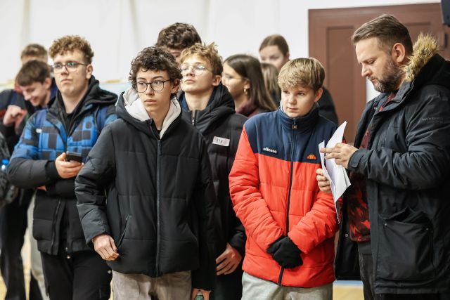 Na zdjęciu znajduje się grupka młodzieży w kurtkach oraz mężczyzna po prawej, który trzyma w ręce kartki.