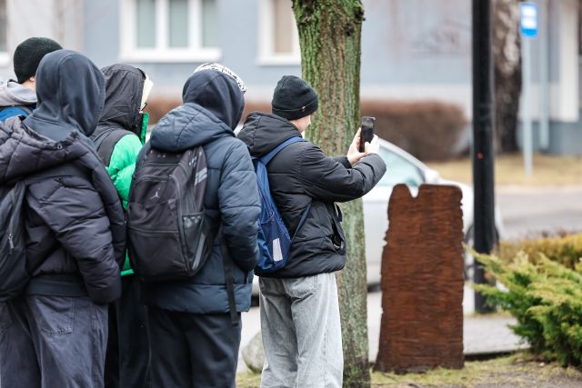 Na zdjęciu znajduje się grupa chłopaków na dworze, jeden z nich ma wyciągnięty telefon przed siebie i robi zdjęcie.