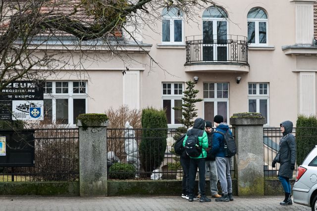 Na zdjęciu znajduje się grupa młodziezy przed budynkiem muzeum i rozwiązują zadania z gry moblinej.