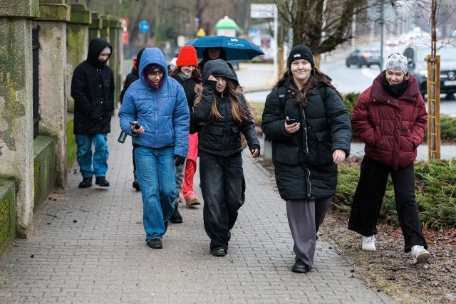 Na zdjęciu znajduje się idąca grupka młodzieży z telefonami, jedna osoba z tyłu ma rozłożony parasol. Grupa znajduje się przy budynku.