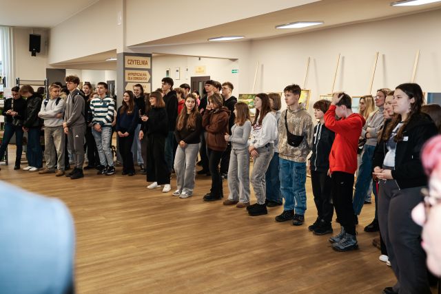 Na zdjęciu znajduje się młodzież stojąca na holu biblioteki. Zdjęcie zostało wykonane od boku.
