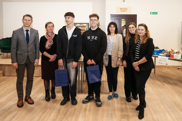 Na zdjęciu znajduje się dwójka nastolatków z upominkami w ręce, z nimi do zdjęcia stoją cztery kobiety oraz jeden mężczyzna. Zdjęcie zostało wykonane na holu.