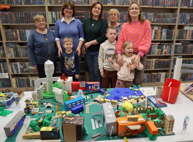 Na zdjęciu znajduje się pięć kobiet oraz trójka dzieci, którzy stoją za wykonaną przez nich makietą miasta leżącą na stole. W tle znajdują się półki z książkami.