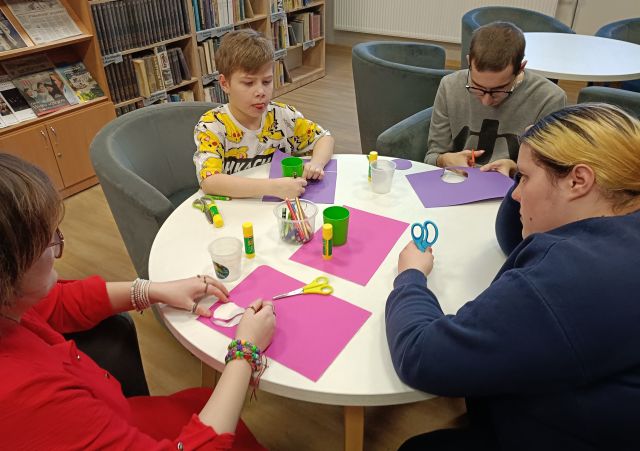 Na zdjęciu znajduje się czwórka młodzieży przy okrągłym stole, na którym wycinają kółka z kolorowych kartek. Na stole znajdują się również kleje, nożyczki i kredki. W tle znajdują się półki z książkami.