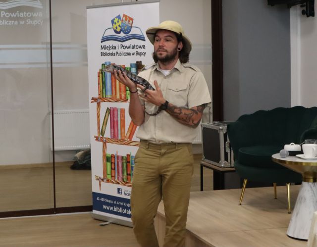 Na zdjęciu znajduje się mężczyzna, który trzyma w rękach jaszczurkę. Za mężczyzną znajduje się baner z książkami i logiem biblioteki, a po prawej stronie mała scena z fotelami i stoliczkiem.