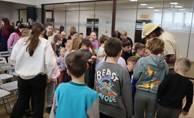 Na zdjęciu znajduje się kolejka dzieci ustawiająca się do mężczyzny w kapeluszu. Po lewej stronie stoją krzesła, a po prawej stronie znajduje się przeszklona ścianka.