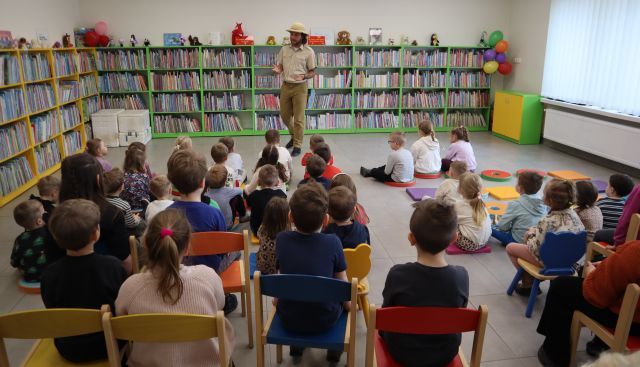 na zdjęciu mężczyzna w stroju podróżnika stojący przed przedszkolakami siedzącymi na podłodze i krzesłach, w tle regały z książkami