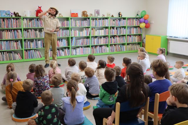 mężczyzna w stroju podróżnika udaje że wkłada do ust robaki, przedn nim na podłodze i krzesełkach siedzą przedszkolaki, w tle regały z książkami