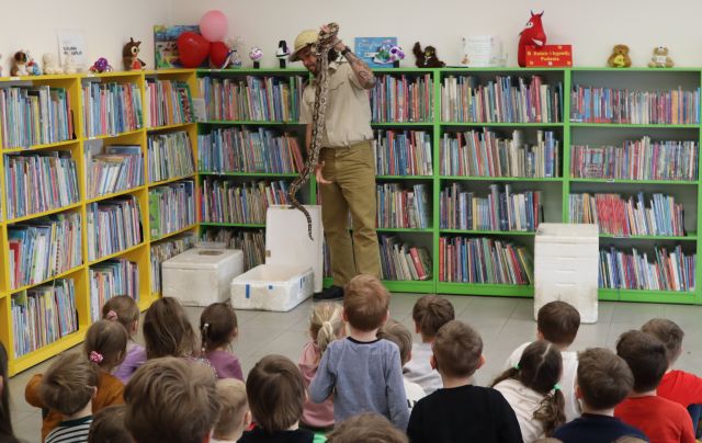 mężczyzna podróżnik wyjmuje z pojemnika węża boa i pokazuje przedszkolakom, w tle regały z książkami