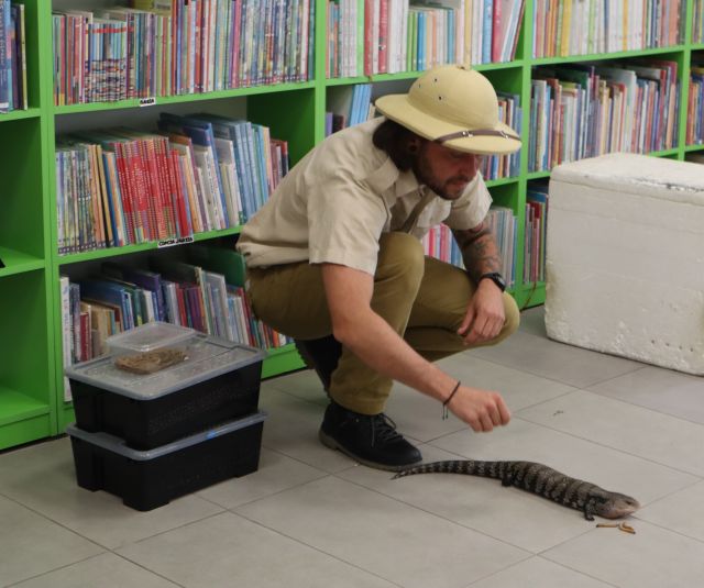 mężczyzna podróżnik kucający przy jaszczurce na podłodze, w tle regały z książkami