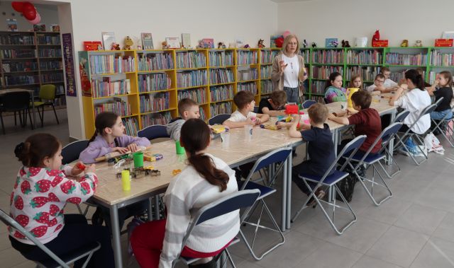 Na zdjęciu znajdują się dzieci przy złączonych stołach, na których znajdują się kubeczki, wałki. Przy stole stoi kobieta, a w tle znajdują się półki z książkami.