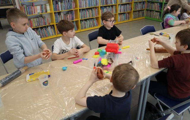 Na zdjęciu znajdują się dzieci przy złączonych stołach, w rękach ugniatają glinę, a na stole leżą wałki i wzory do pierników. W tle znajdują się półki z książkami.