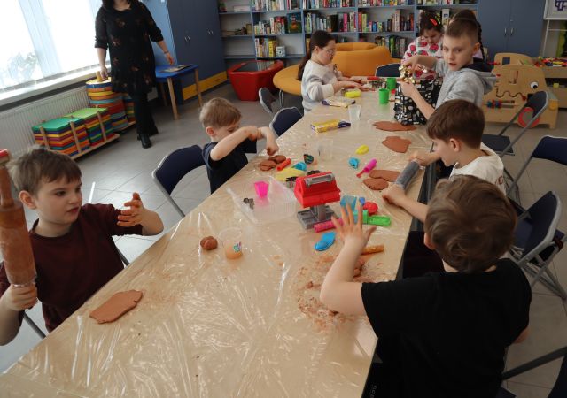 Na zdjęciu znajdują się dzieci przy złączonych stolikach i wałkują glinę, na stole znajdują się również wzory do pierników. W tle znajdują się półki, oraz kobieta.