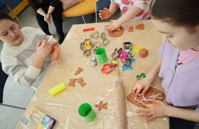 Na zdjęciu znajdują się trzy dziewczynki przy stole, które wycinają wzory z gliny, na stole przed nimi leży wałek, kubek i wzory do pierników.