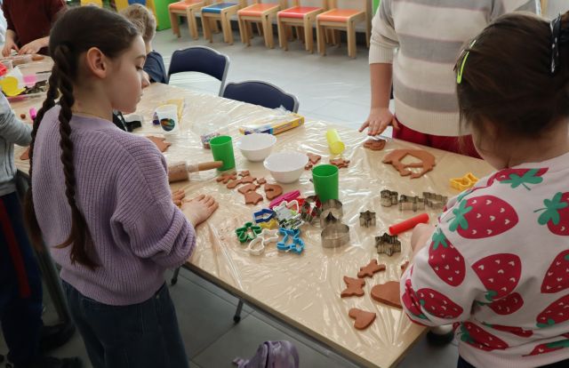 Na zdjęciu znajdują się trzy dziewczynki stojące przy złączonych stołach przy których tworzą pierniki z gliny, za pomocą wzorów do pierników. Zdjęcie zostało wykonane od boku.