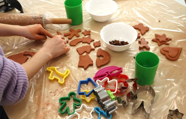 Na zdjęciu znajduje się owinięty folią stół, na którym znajdują się pierniki z gliny, foremki do pierników, kubeczki z wodą i miseczki z ozdobami, na zdjęciu znajdują się również po prawej stronie ręce dziewczynki, która układa z gliny.