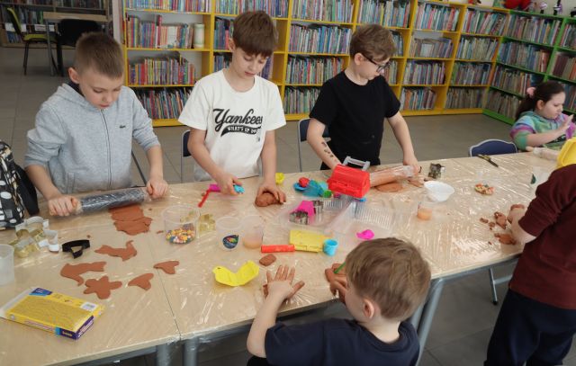 Na zdjęciu znajduje się czwórka chłopców przy złączonych stołach, trzech z nich stoi i robią pierniki z gliny, na stole znajdują się wałki i formy do pierników. W tle znajdują się półki z książkami.