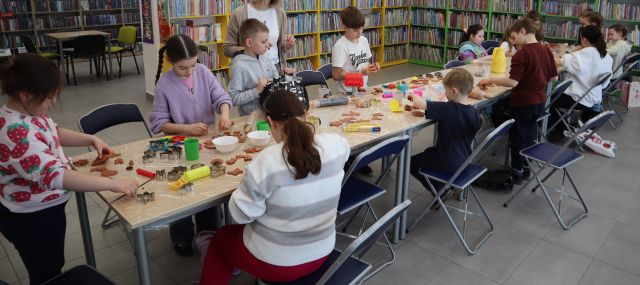 Na zdjęciu znajdują się dzieci przy złączonych stołach, część siedzi, a część stoi. Na stolikach znajduje się glina, foremki do pierników z których uczestnicy tworzą pierniki z gliny. W tle znajdują się półki z książkami.