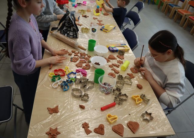 Na zdjęciu znajduje się stół, na którym znajują się foremki do pierników, ozdoby, wałki i pierniki z gliny, które są ozdabiane przez dwie dziewczynki przy stole. Zdjęcie zostało wykonane od góry.