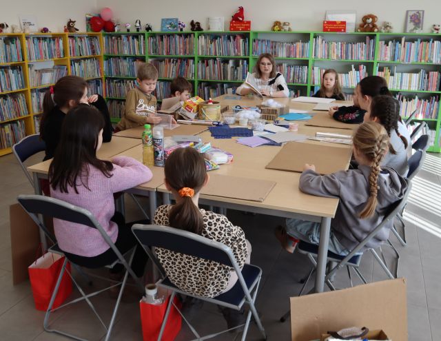 Na zdjęciu znajdują się dzieci przy złączonych w prostokąt stolikach, na których znajdują się kartony, kleje, kartki. Przy stoliku siedzi również kobieta, która ma przed sobą książkę. W tle znajdują się półki z książkami.