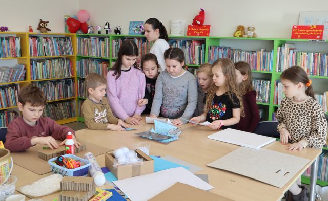 Na zdjęciu znajdują się dzieci na rogu złączonych stołów i próbują ułożyć puzzle. Na stole przed nimi znajdują się kartony, kleje. W tle znajdują się półki z książkami.