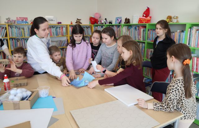 Na zdjęciu znajdują się dzieci przy stole i sięgają po kawałki puzzli. Na stole znajdują się kartony, kleje. W tle znajdują się półki z książkami.