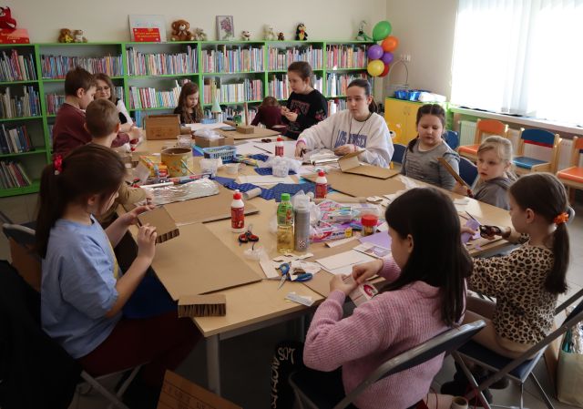 Na zdjęciu znajduje się jedenaścioro dzieci i jedna kobieta przy złączonych w prostokąt stołach i tworzą pałac Królowej Śniegu z materiałów znajdujących się na stole. W tle znajdują się półki z książkami.