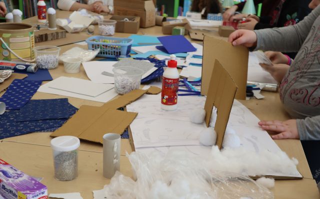 Na zdjęciu znajdują się złączone stoliki z kolorowymi kartkami, kartonikami, klejem. Po prawej stronie na stoliku dziewczynka klei karton tworząc przestrzenny pałac.