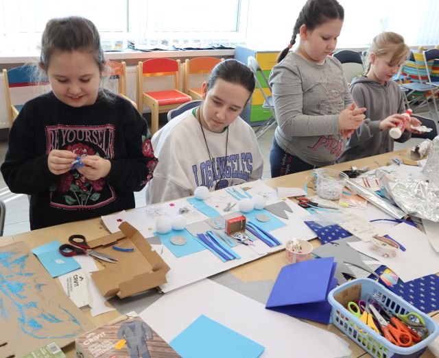 Na zdjęciu znajduje się cztery dziewczynki, trzy z nich stoją przy złączonych stołach i tworzą swój pałac Królowej Śniegu z kolorowych kartek, kartonów znajdujących się na stole. W tle znajdują się okna.