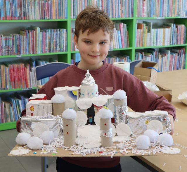 Na zdjęciu znajduje się chłopiec trzymający przed sobą duży pałac wykonany na kartonie, z rolek po papierze, pudełek, wacików, waty i folii aluminiowej. W tle znajdują się półki z książkami.
