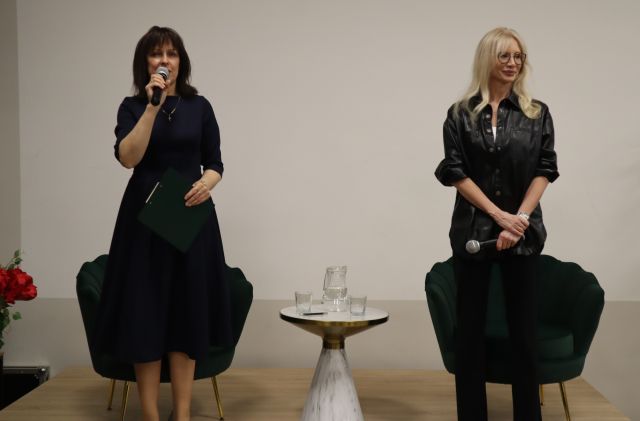 Na zdjęciu znajdują się dwie kobiety na scenie, jedna trzyma przy ustach mikrofon i teczkę w drugiej ręce, między nimi znajduje się stolik.